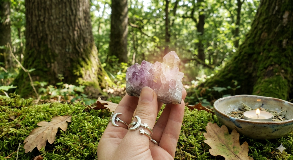 A hand wearing crescent moon and star rings gently holds a cluster of amethyst and clear quartz crystals. The crystals have a subtle, warm glow. The background is a lush, vibrant green forest floor with dappled sunlight filtering through the trees, creating a mystical atmosphere. A small offering bowl with dried herbs and a flickering tealight is visible.